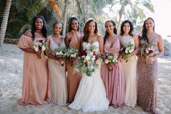 Beautiful bride in white dress with sweetheart neckline stands on the beach with her bridesmaids and holds moody wedding bouquets at her destination wedding in Sayulita, Mexico