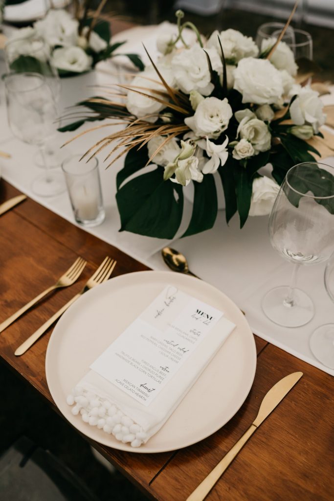 all-white floral arrangments with hints of tropical leaves on the large wooden farmtables with gold utencils, neutral colored ceramic plates and crystal glasswear